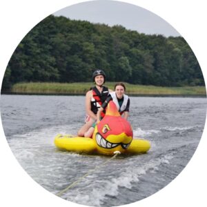 Dwie osoby na zabawce przypominającej rekina jest holowana za motorówką. Jest to wyjazd integracyjny dla klas pierwszych nad jezioro.